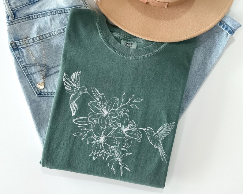 May include: A green t-shirt with a white hummingbird and floral design. The design features a hummingbird flying near a cluster of lilies.
