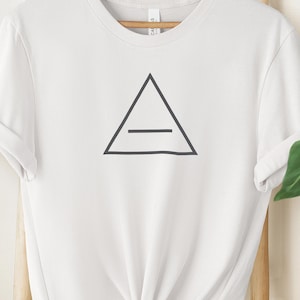 Puede incluir: Camiseta blanca con un diseño geométrico negro de un triángulo con una línea horizontal. La camiseta tiene mangas cortas y cuello redondo. La tela parece suave y cómoda.