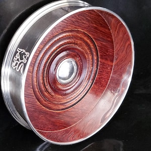 Peut inclure: Un dessous de verre rond avec un bord argenté et un intérieur en bois brun rougeâtre foncé. Le grain du bois est visible en cercles concentriques. Un emblème argenté est sur le côté du dessous de verre.