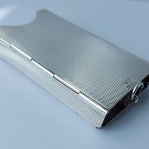 May include: A rectangular, silver-colored cigarette case with a textured, cross-hatched pattern. The case has a curved top and a hinged lid. A small metal ring is attached to the bottom corner.