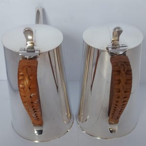 May include: Two silver-plated jugs with matching lids and brown woven handles. The jugs have a cylindrical shape and a shiny finish. The handles are intricately woven, adding a textural contrast to the smooth metal.
