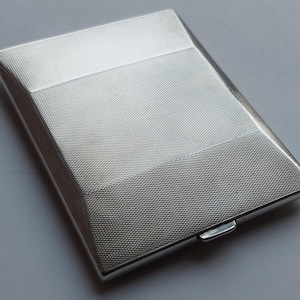 May include: A rectangular silver-toned metal case featuring a fine engine-turned crosshatch texture across its surface. The object has a smooth, polished border and a small, protruding latch mechanism on the side for opening and closing the container.