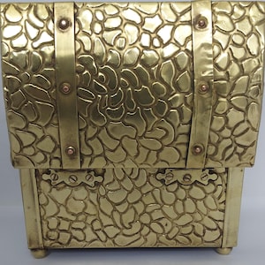Puede incluir: Una caja rectangular de latón con una superficie texturizada similar a guijarros. La caja presenta dos correas verticales de latón aseguradas con remaches de color cobre y dos bisagras horizontales en el panel frontal. Se apoya sobre cuatro pequeñas patas.
