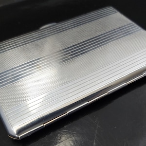 May include: A rectangular, silver cigarette case with a textured surface and horizontal striped design. The case has a hinged lid and a polished finish. The design is simple and elegant, reflecting light.