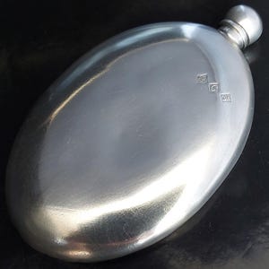Puede incluir: Una petaca de metal de color plateado con forma ovalada, superficie lisa y reflectante y un tapón de rosca redondeado. El cuerpo de la petaca presenta tres pequeños sellos distintivos cerca de la parte superior, indicando su origen.