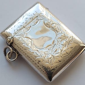 May include: A rectangular, silver-colored antique box with intricate floral and leaf engravings. The box has a hinged lid and a small ring attached to the side. The surface is reflective, and the design is symmetrical.
