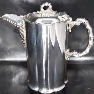May include: A polished silver-toned coffee pot featuring a cylindrical body with vertical fluted panels, a curved spout with ornate leaf-like detailing, a decorative handle with a segmented design, and a lid topped with a sculpted floral finial.