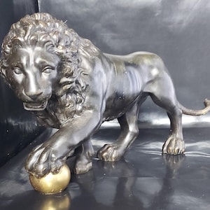 May include: A bronze-colored lion statue with a detailed mane and muscular build. The lion is posed with one paw resting on a small, golden sphere. The sculpture has a dark, antique finish and is set against a dark background.