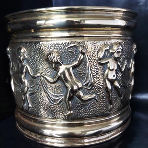May include: A cylindrical brass planter featuring a raised relief design of dancing cherubs encircling the exterior. The surface has a textured background behind the figures, with smooth, polished brass rims at the top and bottom edges.