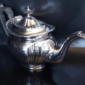 Puede incluir: Una tetera de plata con un acabado brillante y pulido. La tetera tiene un cuerpo redondo con un diseño decorativo y una tapa con un pomo en la parte superior.