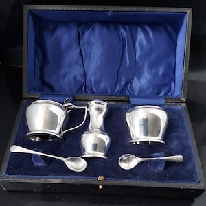 Op de afbeelding: Een zilveren theeservies met een melkkan, suikerpot en twee lepels, gepresenteerd in een zwarte doos met een blauwe fluwelen voering. De set bevat een kleine kan en een vaasvormige container.