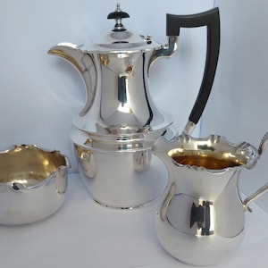 May include: A silver tea set comprising a teapot, creamer jug, and sugar bowl. The teapot features a black handle and a decorative top. The creamer and sugar bowl have gold interiors. The set is highly polished.