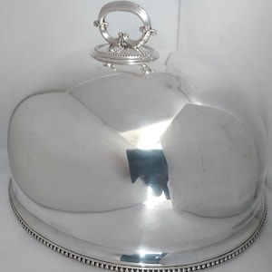 May include: A polished silver-tone dome-shaped food cover with a decorative handle on top and a beaded edge along the base. The reflective metal surface features a large opening in the front, revealing a white interior and a dark, angular object inside.