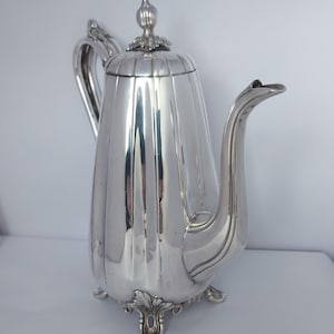May include: A polished silver-toned coffee pot featuring a tall, vertically fluted body, a curved handle, and a long, elegant spout. The lid is topped with a decorative finial, and the base rests on four ornate, scrolled feet with leaf details.