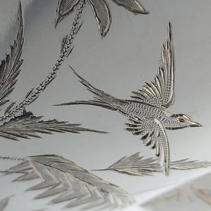 Può includere: Una superficie metallica color argento lucido con un intricato disegno inciso di un uccello in volo con le ali spiegate, circondato da dettagliati elementi botanici tra cui foglie lunghe e sottili e un alto stelo di pianta strutturato.