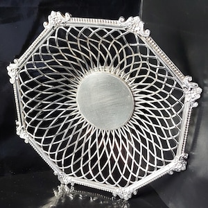Puede incluir: Una cesta octogonal plateada con un diseño calado. La cesta presenta un círculo central sólido y un borde decorativo y ornamentado. El patrón calado crea un efecto de enrejado, y los bordes están detallados con pequeños elementos repetitivos.