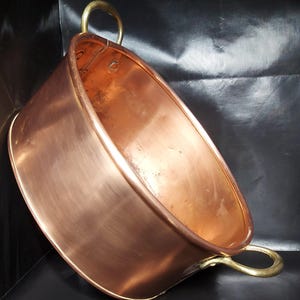 May include: A large, round, polished copper pot with a smooth, reflective surface. The vessel features two sturdy, curved brass handles attached to opposite sides near the top rim. The interior is also copper, showing a warm, metallic finish.