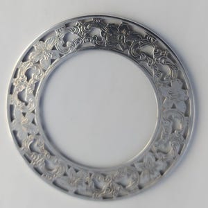 Op de afbeelding: Een zilverkleurige, cirkelvormige decoratieve plaat met een ingewikkeld bloemen- en krulwerkontwerp. De plaat heeft een brede rand met uitgesneden patronen van bloemen en bladeren, waardoor een opengewerkt effect ontstaat. De binnenste cirkel is effen.