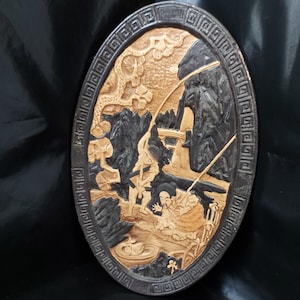 Puede incluir: Un relieve ovalado de madera que representa una detallada escena de paisaje asiático. La obra de arte presenta figuras, montañas y árboles, con un borde oscuro y tallas intrincadas. La paleta de colores incluye tonos de marrón y negro.
