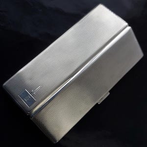 May include: A silver cigarette case with a textured surface and a small engraved logo. The case has a hinged lid and a clasp.