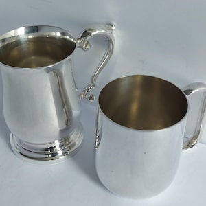 Op de afbeelding: Twee zilverkleurige mokken. Eén mok heeft een gebogen handvat en een bredere basis, de andere is een kleinere, cilindrische mok met een eenvoudig handvat. Beide mokken hebben een glanzend, reflecterend oppervlak, wat suggereert dat ze van metaal zijn.