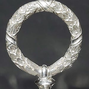 May include: A silver-coloured metal handle featuring a circular ring top with a textured, embossed leaf and vine pattern. The ring is attached to a smooth, tapered base, showcasing a metallic finish with intricate detailing throughout the surface.
