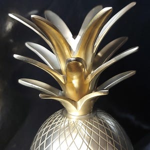 May include: A metallic pineapple sculpture featuring a polished gold-toned top with pointed, layered leaves and a textured, cross-hatched silver-toned base. The object displays a reflective finish and a symmetrical, stylized design of the fruit.