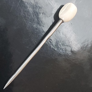 Könnte beinhalten: Eine lange, silberfarbene Hutnadel mit einem cremefarbenen, ovalen Kopf. Die Nadel ist an einem Ende spitz und hat eine glatte, polierte Oberfläche. Die Hutnadel wird wahrscheinlich zum Befestigen von Hüten oder anderen Kopfbedeckungen verwendet.