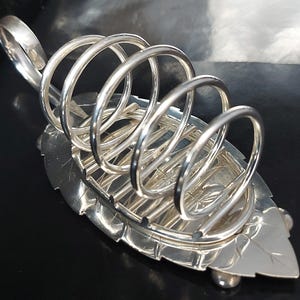 Könnte beinhalten: Ein silberfarbener Toastständer mit einem blattförmigen Sockel und einem Griff. Der Ständer hat eine Reihe von gebogenen, kreisförmigen Haltern für Toastscheiben. Das Design ist verziert und elegant, geeignet zum Servieren von Toast.