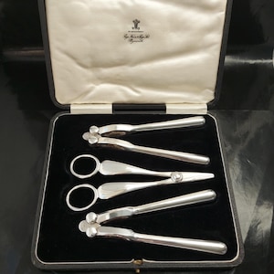 May include: A set of silver-toned nutcrackers and scissors-style tools, presented in a black box with a cream-colored interior. The tools are arranged neatly on a black velvet surface. The box lid has text and a crest.