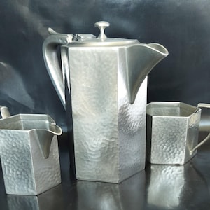 May include: A three-piece silver-toned metal tea set featuring a large hexagonal teapot with a lid, a matching creamer, and a sugar bowl. Each piece has a distinct hammered texture on the exterior surfaces, angular handles, and a polished finish.
