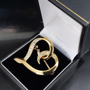 May include: A gold-toned heart-shaped brooch with clear crystal accents, displayed in a black jewelry box with a white interior. The brooch features an intertwined design and a pin closure.