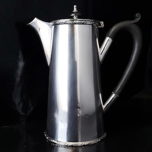 Puede incluir: Una cafetera plateada con un asa negra y un borde decorativo. La cafetera tiene un pico largo y una tapa con un pequeño adorno. El fondo es negro.