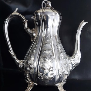 Puede incluir: Una cafetera antigua de color plateado con intrincados grabados florales. La cafetera presenta un pico curvo, un asa grande y una tapa decorativa. Se apoya sobre tres pequeños pies y se encuentra sobre un fondo oscuro.