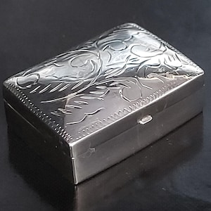 May include: A rectangular silver-colored metal box with a hinged lid. The top surface features an intricate engraved floral pattern with swirling lines and leaves, bordered by a decorative notched edge along the perimeter of the lid.
