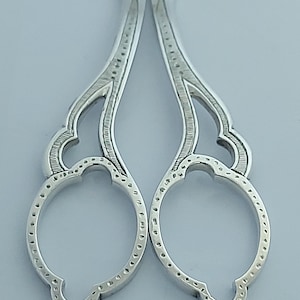 May include: A pair of silver-toned metal scissors featuring ornate, symmetrical handles with intricate etched line patterns and circular finger loops. The design includes decorative cutouts and a textured finish along the edges of the handles.