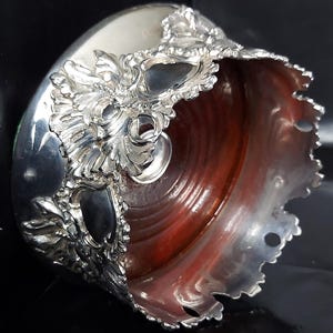 Puede incluir: Un cuenco plateado y ornamentado con un detallado diseño floral alrededor del borde. El interior es de un rico color marrón rojizo. El cuenco tiene un borde festoneado y una pequeña abertura circular en la base.