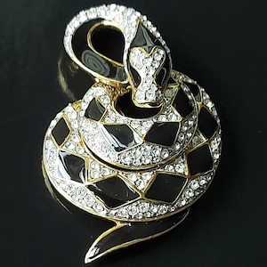Peut inclure: Broche en forme de serpent enroulé, avec une base en métal doré, des accents d'émail noir et des strass transparents étincelants. La tête du serpent est relevée, et le corps est orné de segments noirs et de strass.
