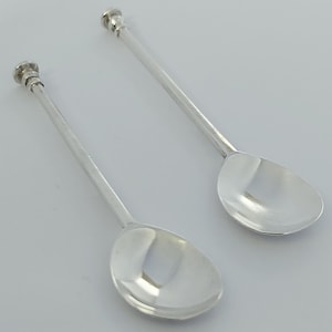 Op de afbeelding: Twee zilverkleurige lepels met lange, slanke stelen en afgeronde, gepolijste kommen. Elke steel is voorzien van een kleine, getrapte, cilindrische knop aan het uiteinde. De lepels hebben een gladde, reflecterende metalen afwerking.