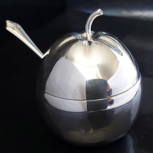 Może przedstawiać: Srebrna cukiernica w kształcie jabłka z pokrywką i pasującą łyżeczką. Miseczka jest wysoko wypolerowana i odbija otoczenie.