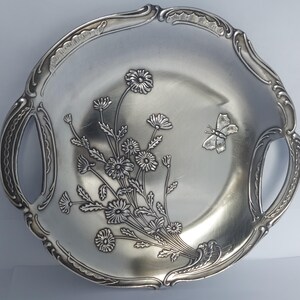Può includere: Un vassoio da portata rotondo in metallo color argento con due manici integrati. Il centro presenta un design in rilievo con un bouquet di margherite e una farfalla. Il bordo ha una finitura decorativa smerlata con dettagli testurizzati.