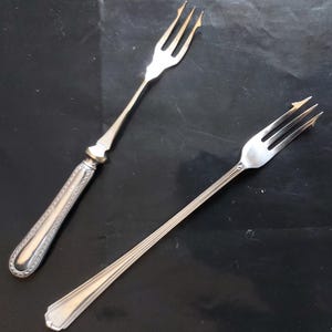 May include: Two silver serving forks with long, thin handles. The forks have three tines each and are designed for serving food.