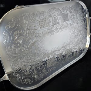 Può includere: Un vassoio da portata color argento con incisioni floreali e a volute ornate. Il vassoio rettangolare ha angoli arrotondati e due manici curvi. Il design dettagliato include un'area ovale centrale e bordi decorativi.