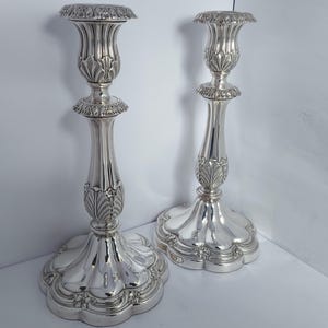 May include: A pair of polished silver-toned metal candlesticks featuring ornate, embossed floral and leaf motifs along the flared bases, the slender stems, and the wide, scalloped candle cups at the top, reflecting light on their smooth surfaces.