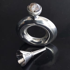 May include: A silver hip flask with a round body and a funnel. The flask has a decorative top with a clear stone. The funnel has the inscription "925" on the side.