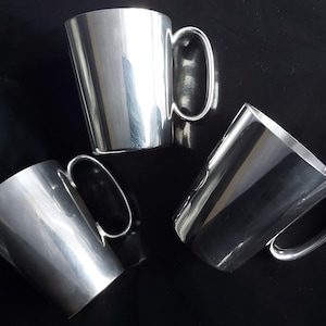 Op de afbeelding: Drie glanzende, zilverkleurige mokken met afgeronde handvatten zijn gerangschikt op een donker oppervlak. De mokken hebben een eenvoudig, elegant ontwerp, dat licht reflecteert en highlights creëert. De handvatten zijn lusvormig en de mokken lijken van metaal te zijn.