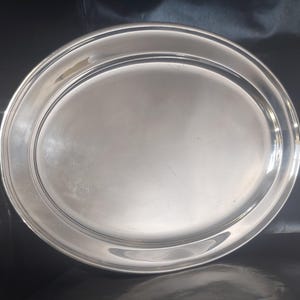May include: A polished, oval silver serving platter. The platter has a smooth, reflective surface with a slightly raised rim and a decorative groove near the edge. The design is simple and elegant, suitable for serving food.