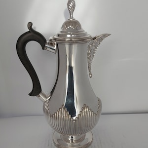 以下が含まれることがあります： 黒い取っ手と装飾的な蓋が付いた銀製のコーヒーポット。ポットはフルーテッドデザインで、丸い底をしています。