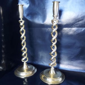 May include: A pair of silver-toned candlesticks. Each candlestick features a twisted stem, resembling a rope, and a circular base. The tops are designed to hold candles. The background is a dark blue fabric.