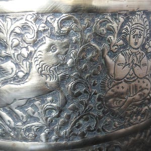 Op de afbeelding: Een metalen kom met een messingkleurige afwerking en ingewikkelde reliëfgravures. Het oppervlak toont een gedetailleerde scène van een gestileerde leeuw in profiel naast een zittende figuur met gevouwen handen, omringd door sierlijke bloemmotieven.